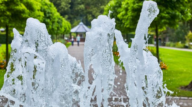 Vordergründig ist in einer Nahaufnahme das sprudelnde Wasser eines Springbrunnens zu sehen. Im Hintergrund ist ein Park mit Pavillon.