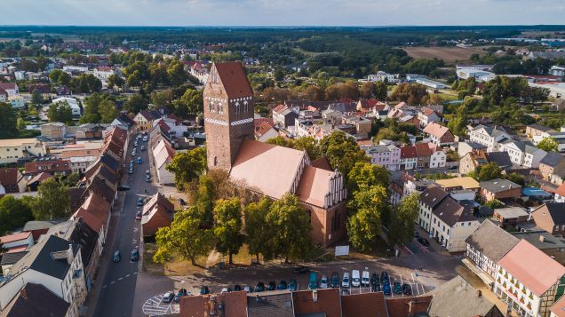 Zu sehen ist ein Dorf aus Vogelperspektive, in dessen Zentrum eine rote Backsteinkirche mit einem Kirchturm steht. Im Hintergrund erstreckt sich ein flachen Waldgebiet.