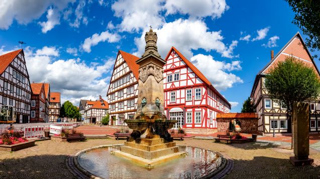 Zu sehen ist ein Dorfplatz mit Altbauten und einem runden Brunnen im Vordergrund. 