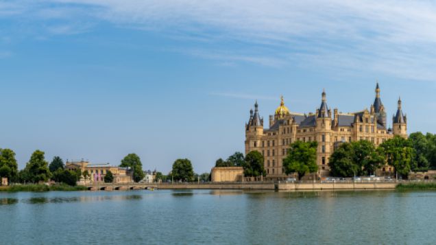 Schloss Schwerin. Quelle: AdobeStock Fotos Im Vordergrund ist ein Gewässer. Dahinter ist das markant gelbe Schloss von Schwerin vor einem blauen Himmel zu sehen.