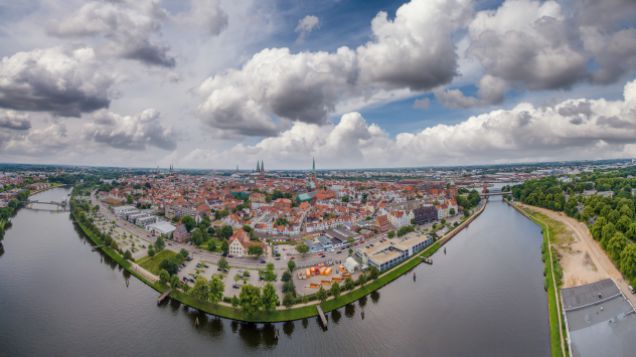 Zu sehen ist die Stadt Lübeck aus Vogelperspektive. Ein Fluß umrahmt die Innenstadt. Diese besteht aus vielen roten backsteinhäusern. Das Bild ist im Weitwinkel aufgenommen.