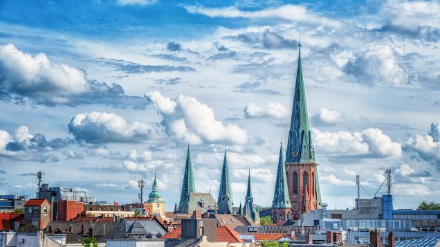 Stadtansicht Oldenburg. Quelle: AdobeStock Fotos Dächer der Stadt Oldenburg. Zu sehen ist ein hoher Turm mit türkisenem Spitzdach. Dieser ist umgeben von weiteren Stadthäusern.