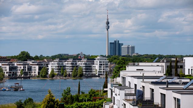 Im Vordergrund sind moderne Mehrfamilienhäuser zu sehen. Dahinter erstreckt sich eine Wasserfläche, die von ähnlichen, modernen Gebäuden gerahmt wird. Im Hintergrund sind hochhäuser und ein Fernsehturm zu sehen. 