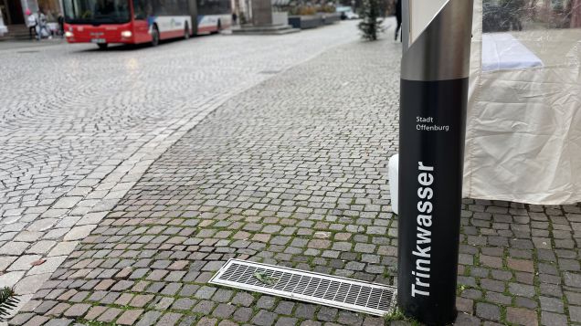 Ein schwarzer Trinkwasserspender steht im Vordergrund an einer Straße aus Pflastersteinen. Im Hintergrund sind Häuser und ein Bus zu sehen.