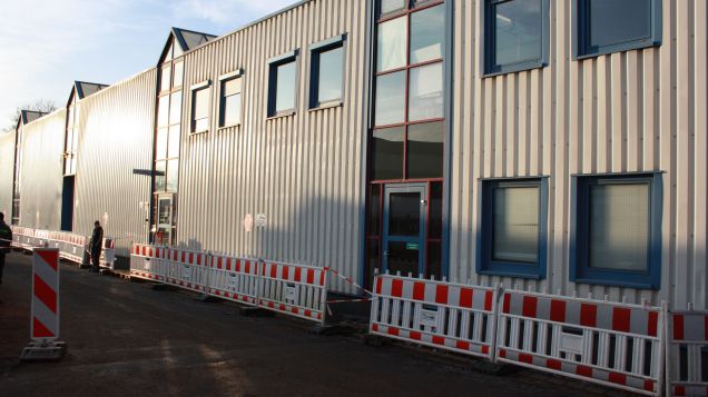 Zu sehen ist eine metallene Fassade der Werkshalle. Diese glänzt in der Sonne. Anstehende Bauarbeiten sind erkennbar an den Bauzäunen am Boden.
