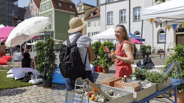 Zwei Bürgerinnen stehen auf einem innterstädtischen Platz vor einem Stand mit kleinen Modellen.