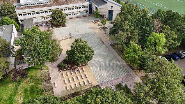 Zu sehen ist ein entsiegelter Schuhol umgeben von Bäumen und Rasen. Hinten ist das Schulgebäude und rechts oben in der Ecke ein Fußballfeld. Auf dem Hof sind zwei kleinere Gruppen von Menschen.
