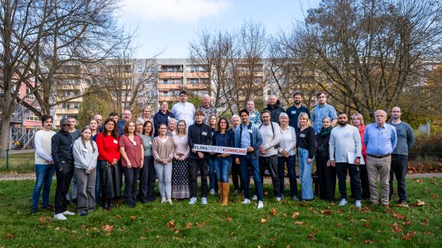 Eine große Gruppe von Menschen steht auf einer Wiese. Die Person in der Mitte hält ein Schild, auf dem steht "Klima trifft Kommune".