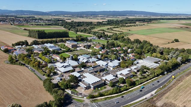 Aus der Vogelperspektive ist ein weitläufiger Campus zu sehen, bestehend aus mehreren Gebäuden. Er ist umringt von Feldern, Waldstreifen und einer Straße. Im Hintergrund sind Berge und davor kleine Orte.