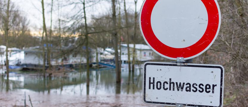 Im Vordergrund ist ein Durchfahrt-Verboten Schild mit der Zusatzinformation Hochwasser. Im Hintergrund ist der Weg mit Wasser überschwemmt.