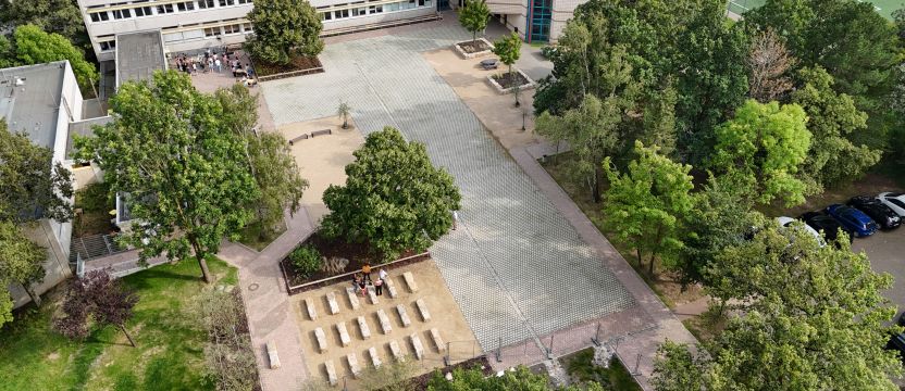 Zu sehen ist ein entsiegelter Schuhol umgeben von Bäumen und Rasen. Hinten ist das Schulgebäude und rechts oben in der Ecke ein Fußballfeld. Auf dem Hof sind zwei kleinere Gruppen von Menschen.
