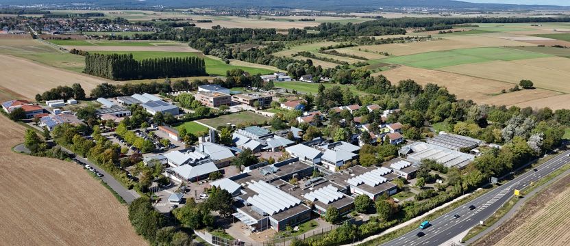 Aus der Vogelperspektive ist ein weitläufiger Campus zu sehen, bestehend aus mehreren Gebäuden. Er ist umringt von Feldern, Waldstreifen und einer Straße. Im Hintergrund sind Berge und davor kleine Orte.
