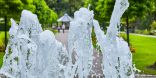 Vordergründig ist in einer Nahaufnahme das sprudelnde Wasser eines Springbrunnens zu sehen. Im Hintergrund ist ein Park mit Pavillon.