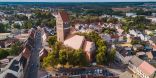 Zu sehen ist ein Dorf aus Vogelperspektive, in dessen Zentrum eine rote Backsteinkirche mit einem Kirchturm steht. Im Hintergrund erstreckt sich ein flachen Waldgebiet.