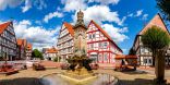 Zu sehen ist ein Dorfplatz mit Altbauten und einem runden Brunnen im Vordergrund. 