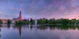 Im Vordergrund ist ein angelegter Teich zu sehen. Im Hintergrund stehen Stadtparkbäume und links im Bild ist ein roter Backsteinturm zu sehen.