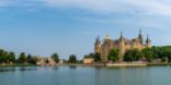 Im Vordergrund ist ein Gewässer. Dahinter ist das markant gelbe Schloss von Schwerin vor einem blauen Himmel zu sehen. 