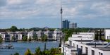 Im Vordergrund sind moderne Mehrfamilienhäuser zu sehen. Dahinter erstreckt sich eine Wasserfläche, die von ähnlichen, modernen Gebäuden gerahmt wird. Im Hintergrund sind hochhäuser und ein Fernsehturm zu sehen. 