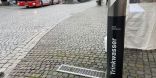 Ein schwarzer Trinkwasserspender steht im Vordergrund an einer Straße aus Pflastersteinen. Im Hintergrund sind Häuser und ein Bus zu sehen.