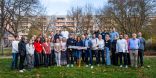 Eine große Gruppe von Menschen steht auf einer Wiese. Die Person in der Mitte hält ein Schild, auf dem steht "Klima trifft Kommune".