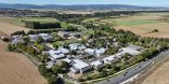Aus der Vogelperspektive ist ein weitläufiger Campus zu sehen, bestehend aus mehreren Gebäuden. Er ist umringt von Feldern, Waldstreifen und einer Straße. Im Hintergrund sind Berge und davor kleine Orte.