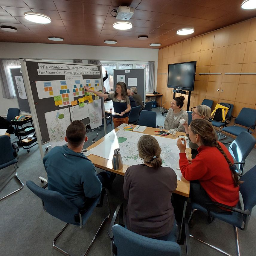 Zu sehen ist ein Innenraum mit einem quadratischen Tisch mittig. Um den Tisch sitzen 5 Personen. Auf dem Tisch liegt eine Karte ausgebreitet. Links neben dem Tisch steht ein Pinnboard, auf das verschiedenfarbige Post-its, Karten und Zettel geheftet sind.