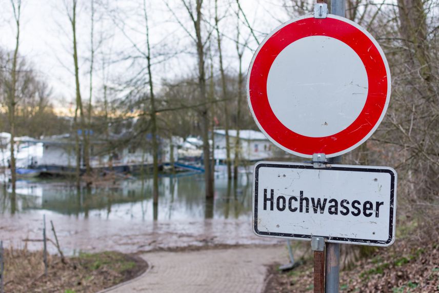 Im Vordergrund ist ein Durchfahrt-Verboten Schild mit der Zusatzinformation Hochwasser. Im Hintergrund ist der Weg mit Wasser überschwemmt.