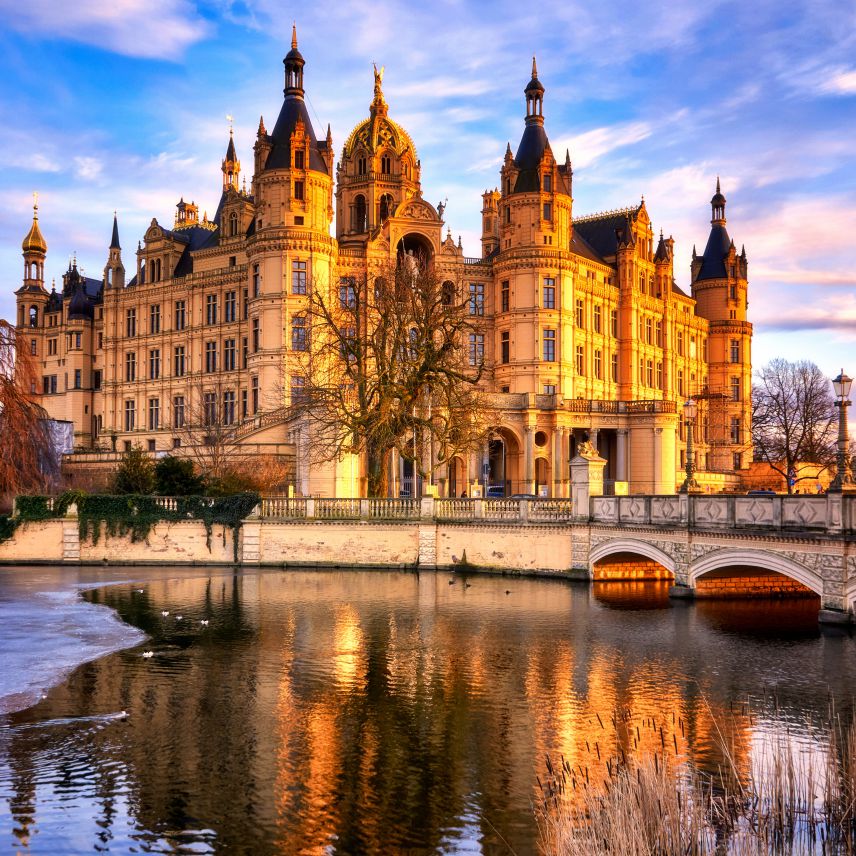 Schloss der Landeshauptstadt Schwerin. Quelle: AdobeStock Fotos Hinter einem Gewässer mit einer Brücke zugänglich steht das Schloss von Schwerin in goldenem Abendsonnenlicht.