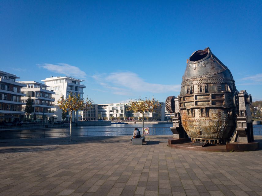 Metall Statue. Quelle: AdobeStock Fotos Im Vordergrund ist ein Platz aus Steinen, auf dem eine metallene Statue eines Fisches steht. Dahinter ist Gewässer erkennbar, das von modernen Gebäudekomplexen gerahmt wird.