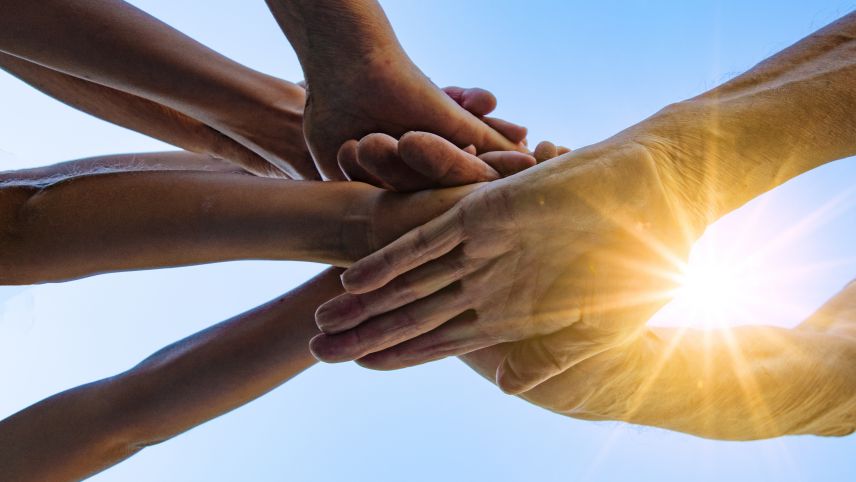 Mehrgenerationen-Arbeit. Quelle: AdobeStock Fotos Zu sehen sind Hände, die sich in der Mitte treffen. Die unterste Hand ist faltig. IM Hintergrund scheint eine Sonne durch Lücken der aufeinander gestapelten Hände.