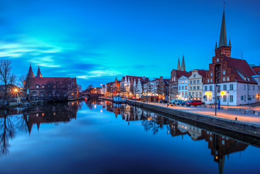 Stadt Lübeck. Quelle: AdobeStock Fotos An einem Gewässer stehen Stadtgebäude. Das Foto ist bei Dämmerung aufgenommen, weswegen alles in ein dunkelblaues Licht getaucht ist.