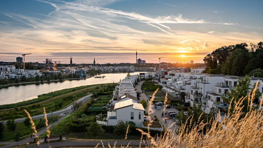 Blick auf Dortmund-Hörde bei Sonnenuntergang. Quelle: AdobeStock Fotos Moderne, helle Gebäude stehen in Reihen parallel zu einem Fluss. Im Hintergrund erstreckt sich die Stadt Dortmund. Das Bild ist bei Sonnenuntergang aufgenommen, wodurch ein goldenes Licht über Szene liegt.