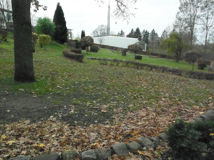 Ein steil abschüssiger Garten ist zu sehen. Im Hintergrund sind Gebäude erkennbar. Hecken, größere Büsche und Bäume sind in dem Gartenstück vorhanden. Im Vordergrund ist eine Steineinfassung und Laub auf dem Boden zu sehen.