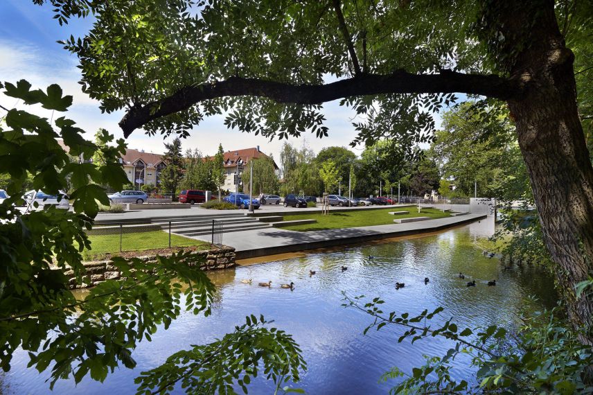 Terassen am Mühlbach. Quelle: Stadt Offenburg Ein Bach eingefasst in Steinufer. Die Betrachterin schaut durch Büsche und Bäume auf die gegenüberliegende Bachseite. Hier sind Terassen mit Sitzmöglichkeiten aus Stein zu sehen. Im Hintergrund ist ein Parkplatz und Häuser sichtbar.