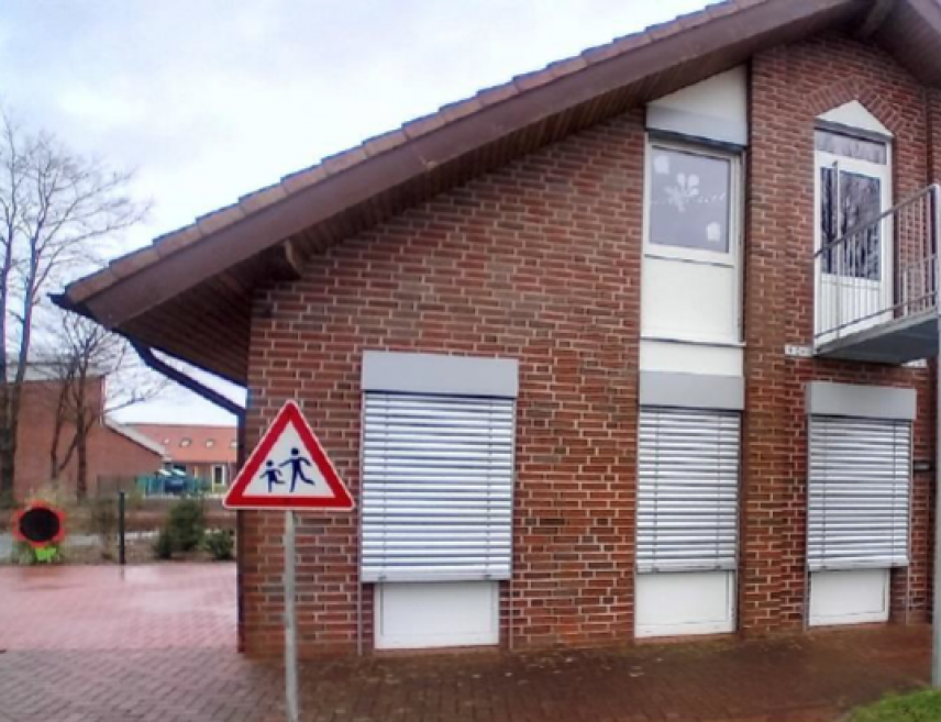 Zu sehen ist eine Backsteinfassade einer Kita mit großen, neuen Fenster. Raffstoffe zur Verschattung sind vor einigen Fenster heruntergelassen.