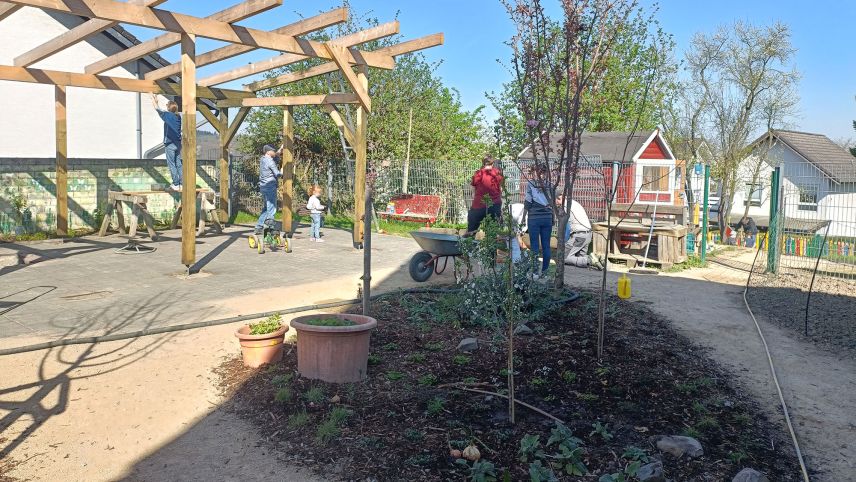 Verschiedene Menschen arbeiten an und neben der Holz-Pergola. Im Vordergrund ist ein begrünter Bereich zu sehen.