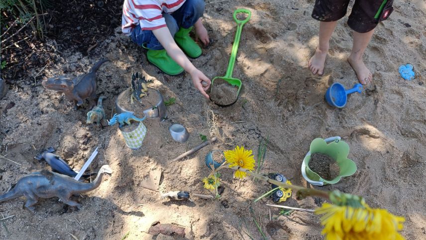 Spielzeuge liegen in einem schattigen Sandkasten. Kinder spielen in dem Sand.
