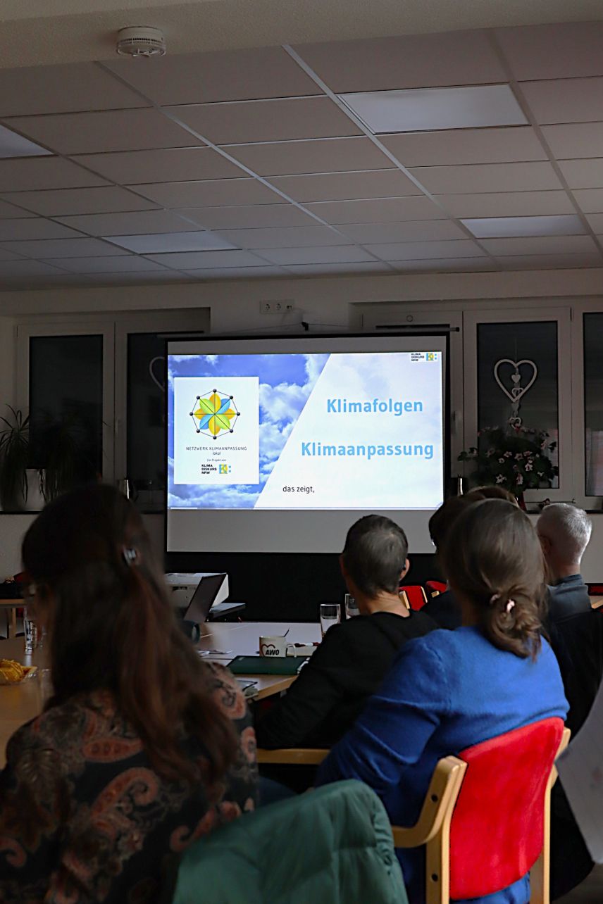 Menschen sitzen an einem Tisch und schauen zu einer Präsentation mit dem Titel Klimafolgen Klimaanpassung.