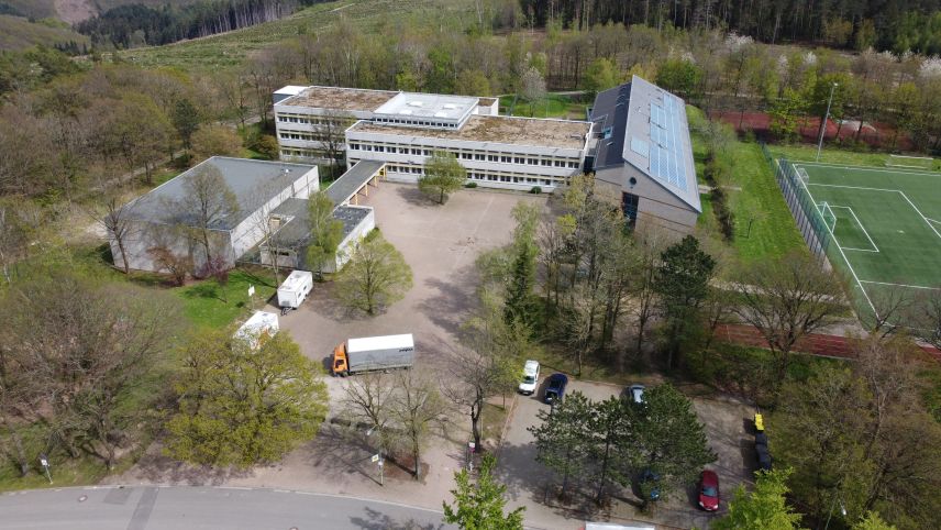 Zu sehen ist ein Schulgelände mit Hof und Schulgebäude. Das Gelände ist umringt von Bäumen. Rechts ist ein Fußballfeld zu sehen. Unten rechts in der Ecke ist ein Parkplatz mit Autos.