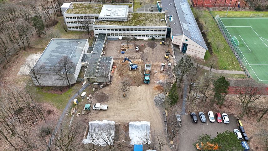Zu sehen ist ein Schulgelände mit Hof und Gebäude. Der Hof ist aufgerissen und mehrere Bagger und LKWs stehen bereit. Rechts sind ein Fußballfeld und ein Parkplatz zu sehen.
