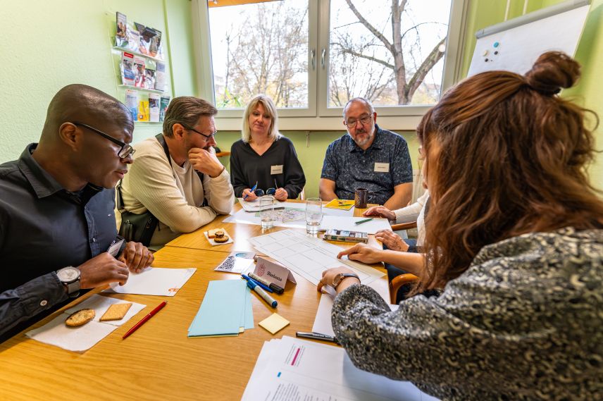 Eine kleine Gruppe von Leuten sitzt an einem Tisch. Es liegen Arbeitsblätter und Stifte auf dem Tisch. Eine Person zeigt auf ein Blatt, die anderen gucken auf die gezeigte Stelle.