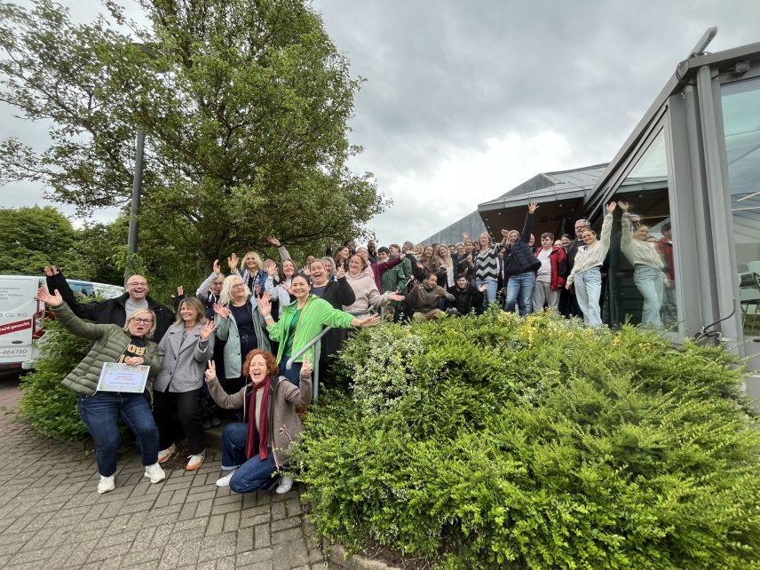 Eine große Gruppe von Leuten ist zu sehen, die alle jubeln oder die Arme in die Höhe reißen. Vorne rechts ist eine Hecke und links ist ein Baum. Rechts im Hintergrund ist ein Haus.